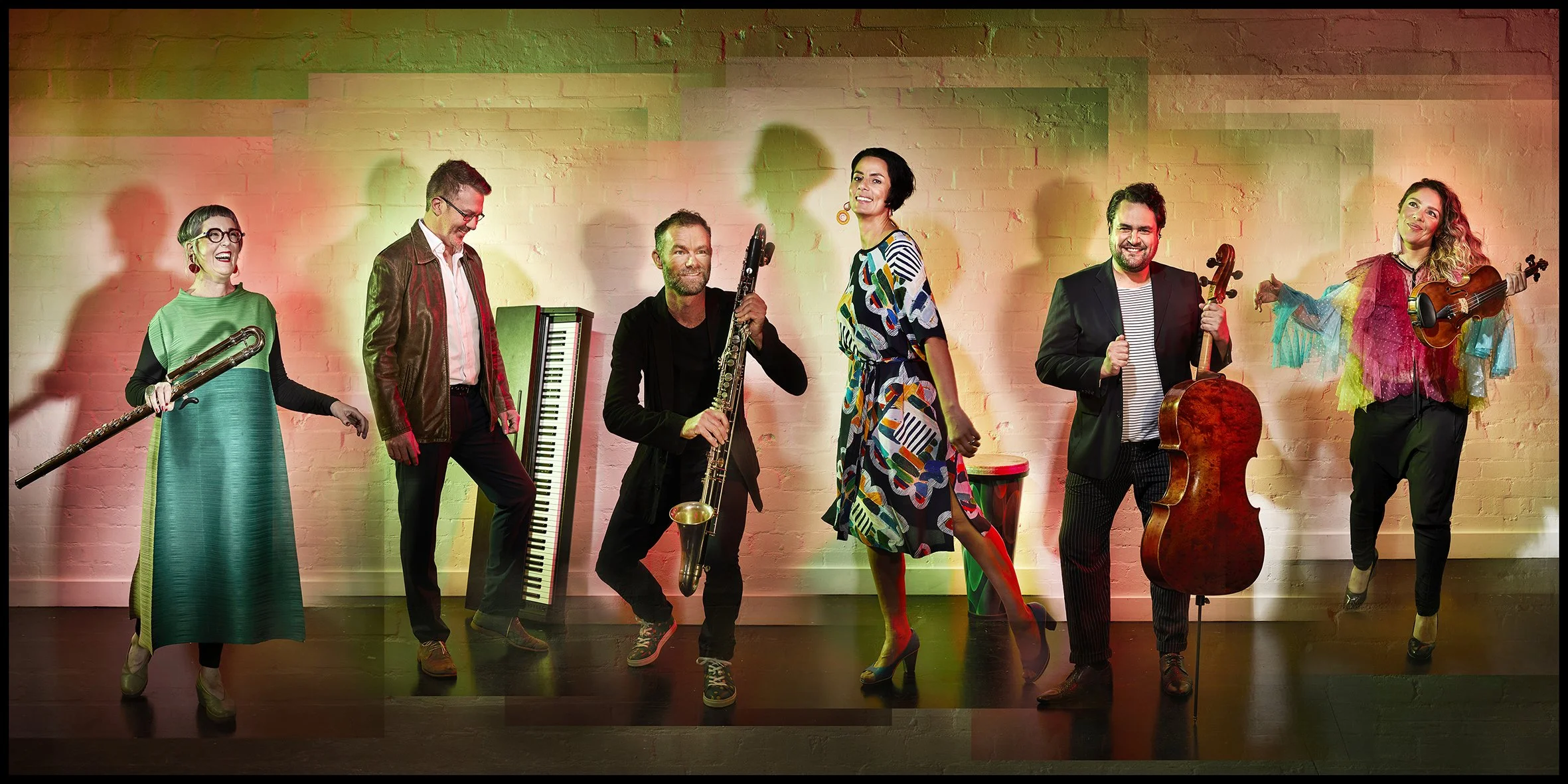 Group of six musicians standing on stage with colorful lighting and a white brick wall background, holding various musical instruments including a flute, keyboard, saxophone, bongos, cello, and violin.