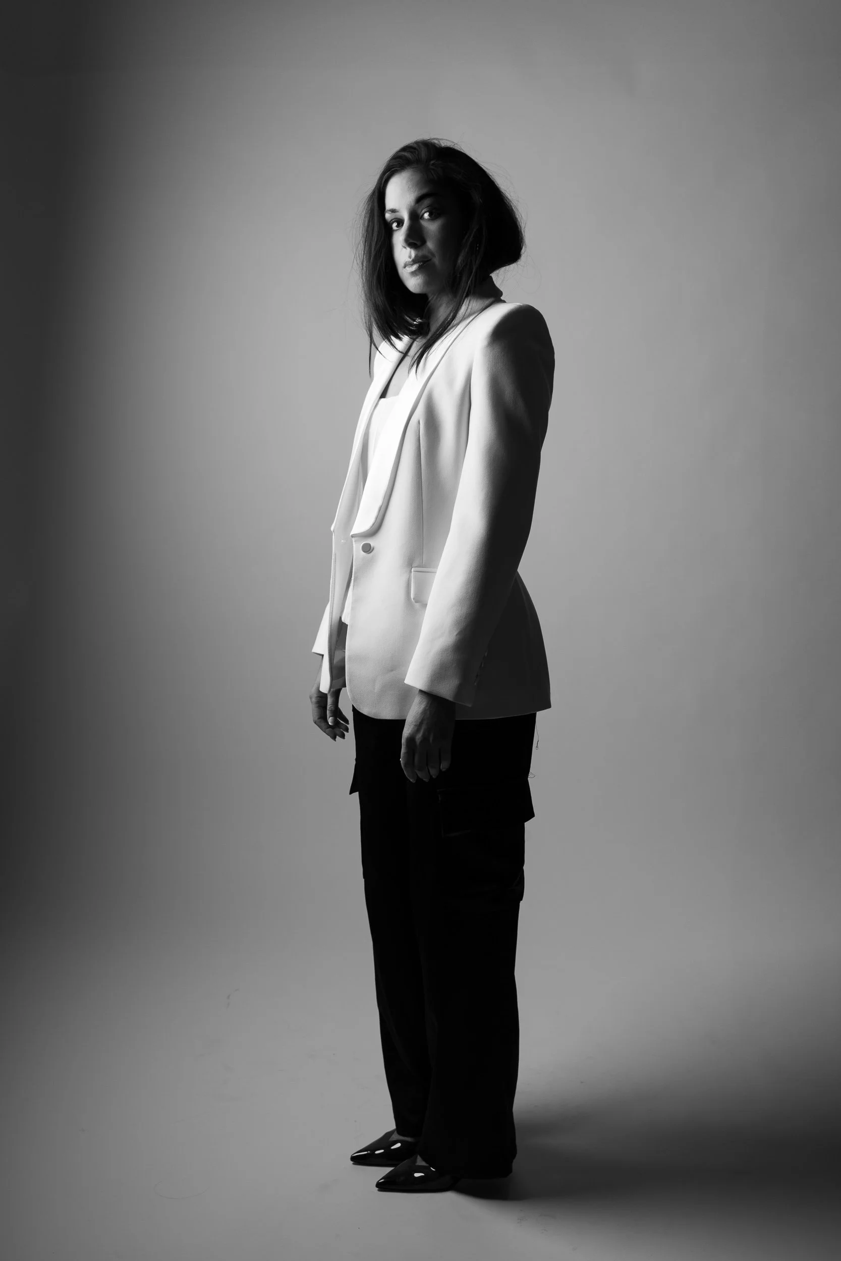 Black and white portrait of a woman standing sideways, wearing a blazer, dark pants, and patterned shoes, with her face turned towards the camera.