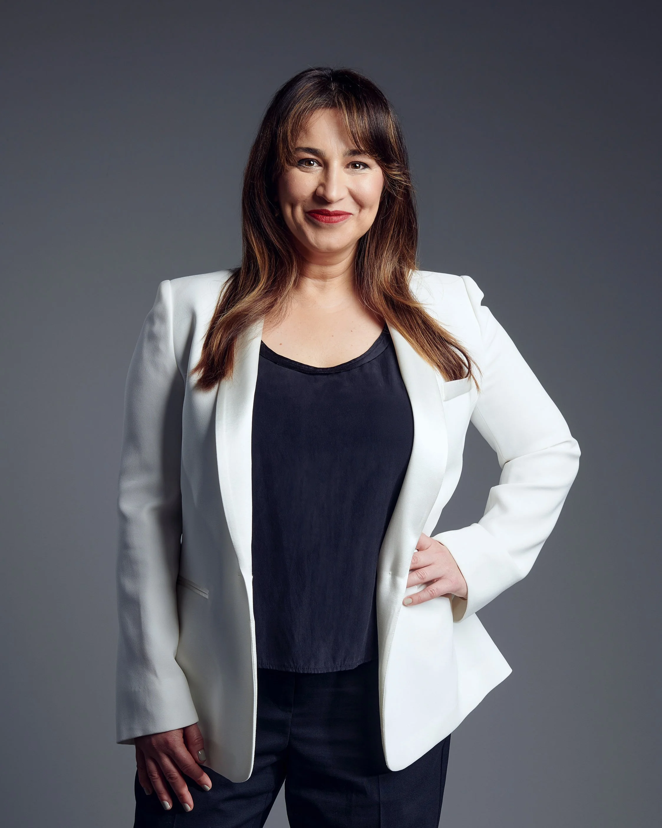 A woman with shoulder-length brown hair in a white blazer and black shirt standing against a gray background, smiling confidently with one hand on her hip.