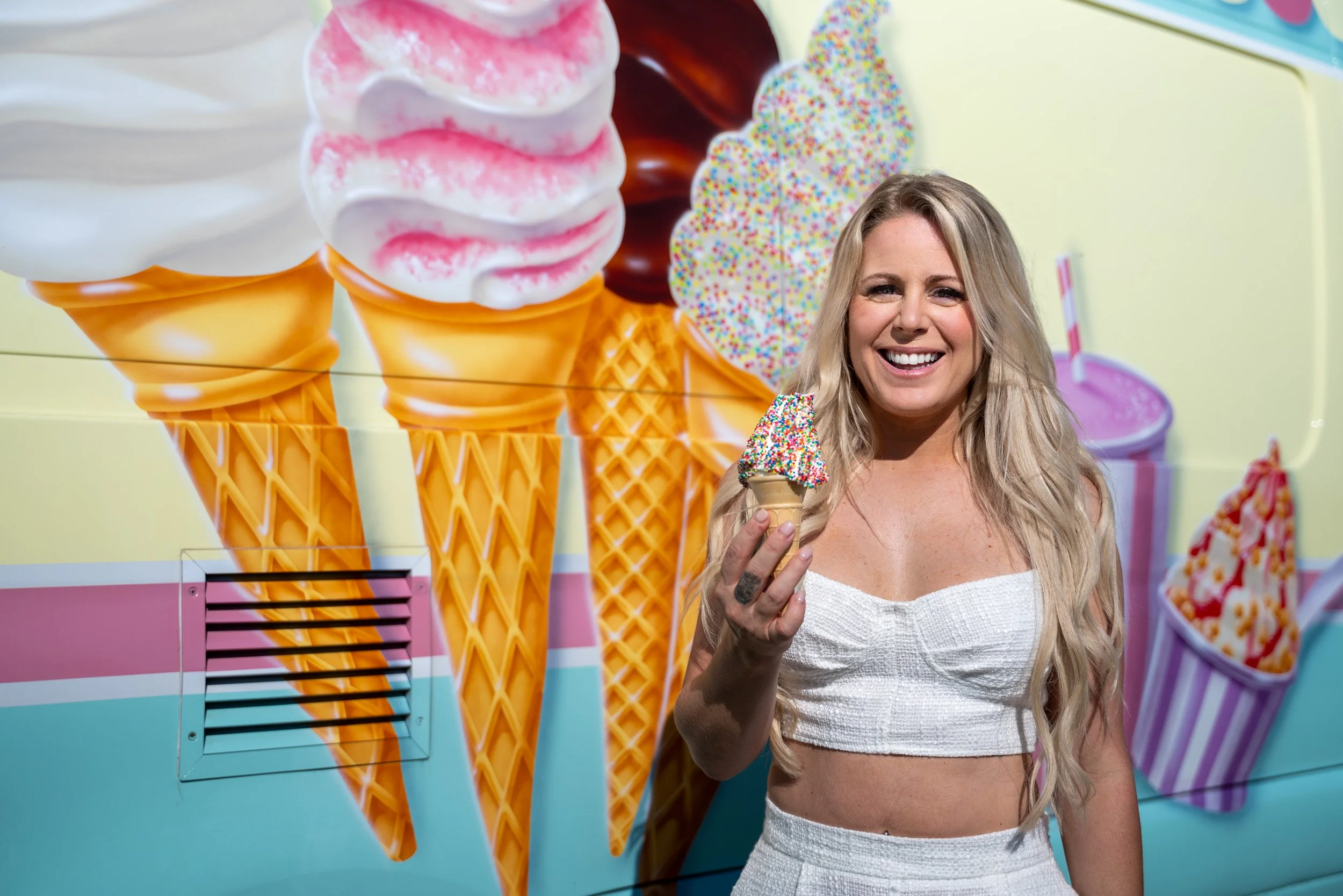 Woman smiling and holding an ice cream cone with colorful sprinkles in front of a large mural of ice cream cones and frozen treats.