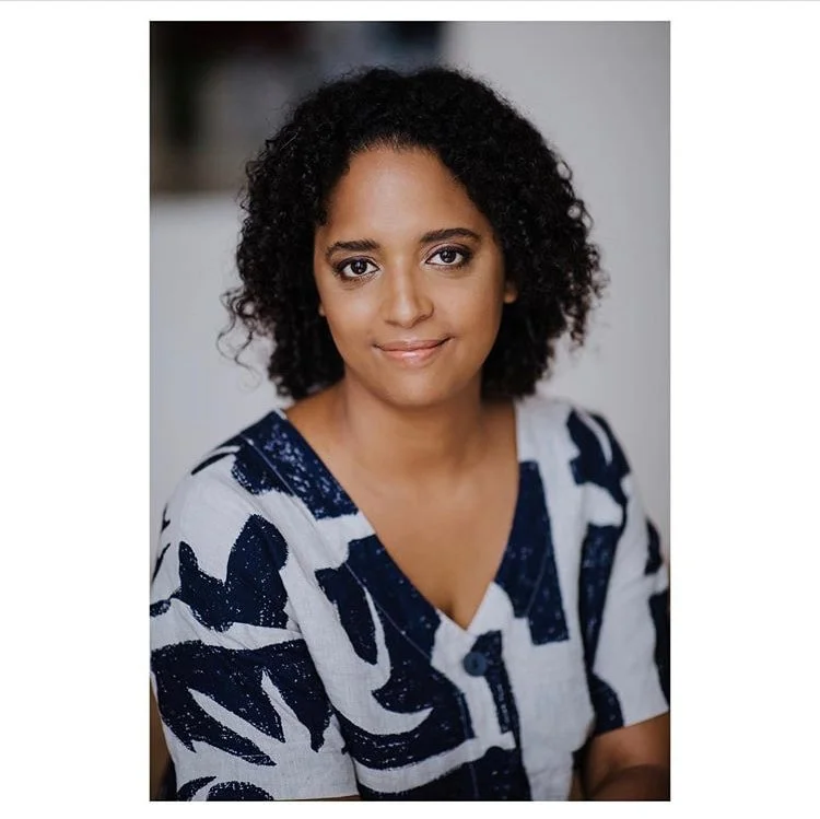 A woman with curly dark hair and medium skin tone, wearing a white and dark blue patterned top, posing indoors with a neutral expression.