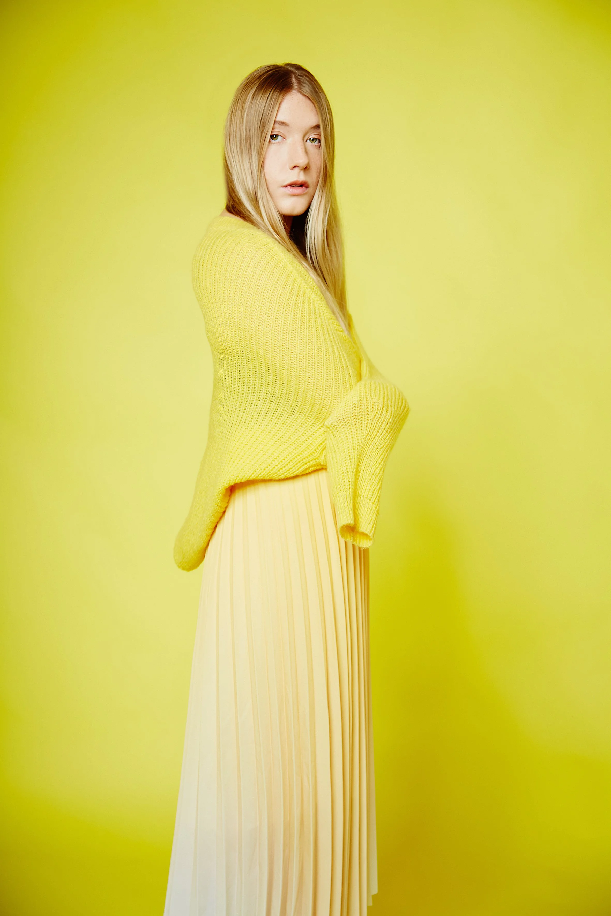 A young woman with long blonde hair and light skin in a yellow sweater and pleated yellow skirt standing against a yellow background.