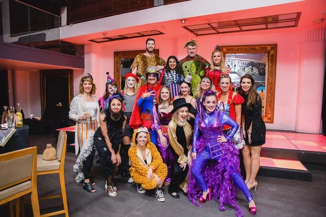 Group of people in colorful costumes at a party, with some wearing hats, masks, and themed outfits, in an indoor setting with pink lighting and framed artwork on the wall.