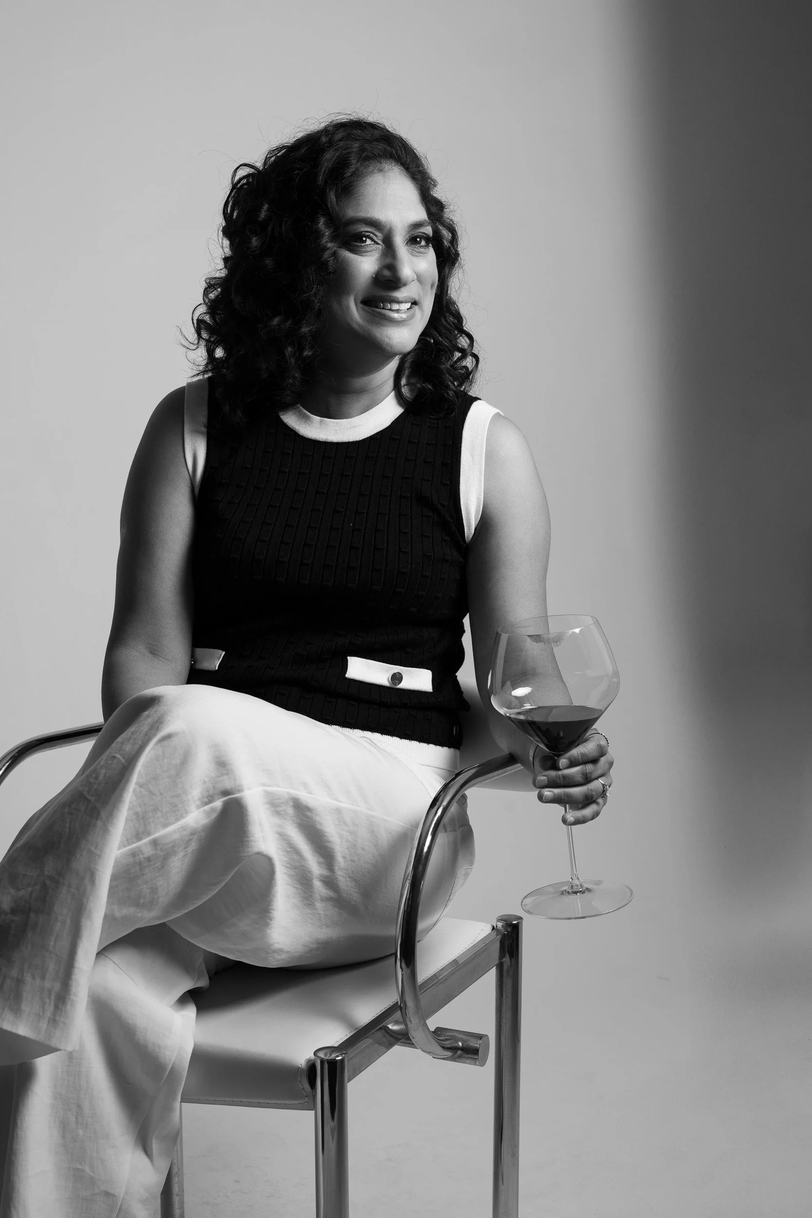 A black and white photo of a woman sitting on a modern chair, holding a glass of red wine, smiling, with curly hair, wearing a sleeveless top and light-colored pants.