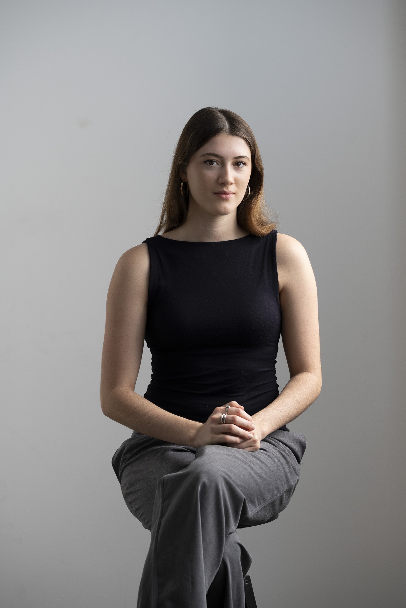 A young woman with long brown hair, wearing a black sleeveless top and gray pants, sitting cross-legged with her hands clasped, against a plain light gray background.