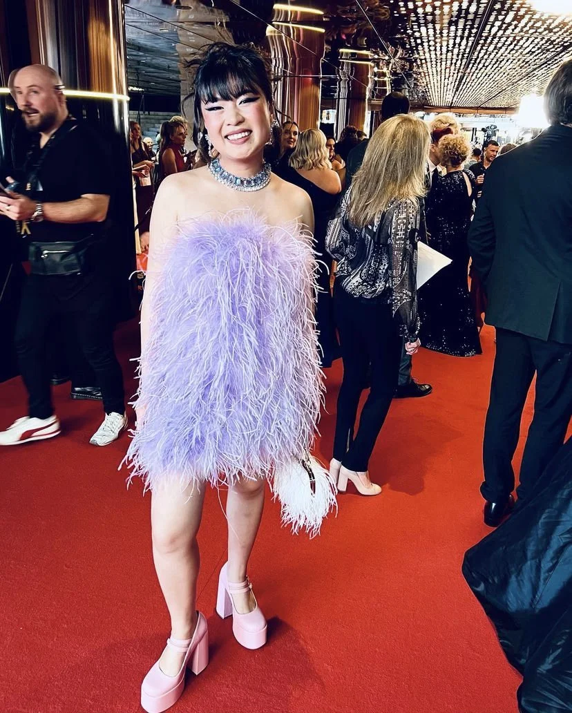 A woman smiling in a purple feathered dress with a choker necklace at a red carpet event, surrounded by people in formal attire.