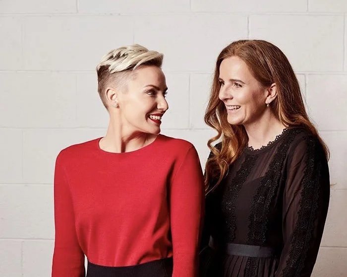 Two women standing close together, smiling and looking at each other, against a white brick wall. one has short, platinum blonde hair and wears a red top, the other has long, red hair and wears a black lace dress.