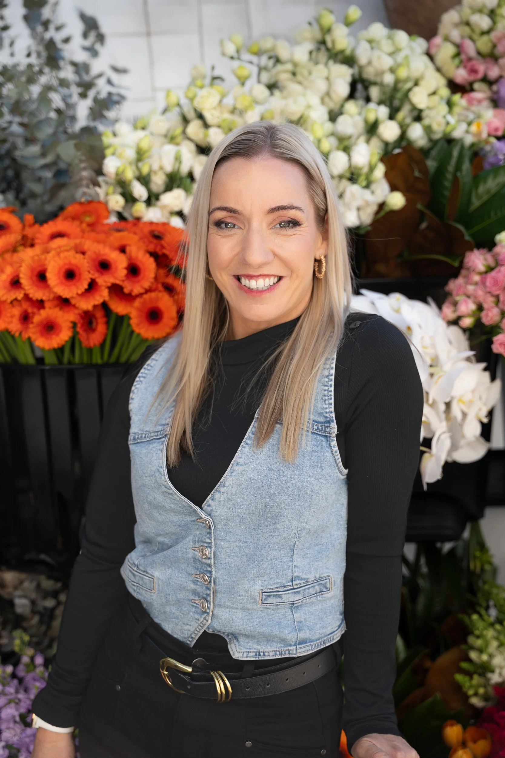 A woman with blonde hair and earrings smiling in front of a display of colorful flowers including white, orange, purple, and pink blossoms.
