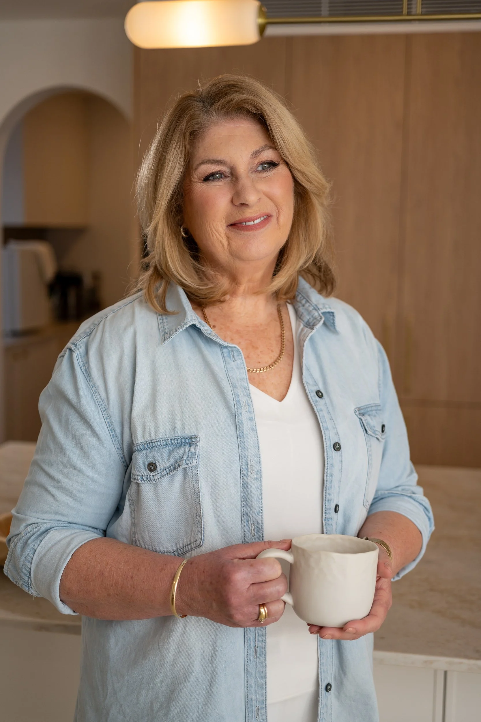 A middle-aged woman with blonde hair smiling, holding a white mug in a modern kitchen.