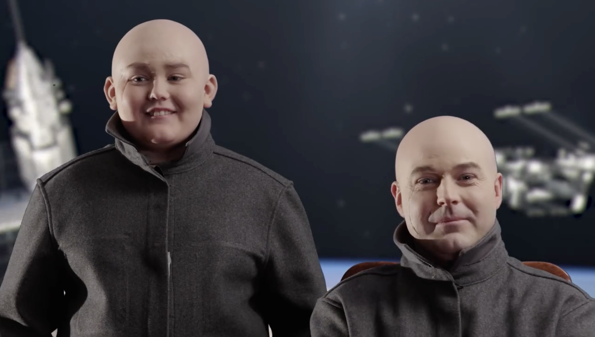 A woman dressed as an alien from the Star Wars franchise, with a bald head and gray uniform, smiling. A man dressed as an alien, with a bald head, gray uniform, and a slight smile, sitting in front of her. Behind them is the backdrop of space, with stars and the Millennium Falcon spaceship from Star Wars.