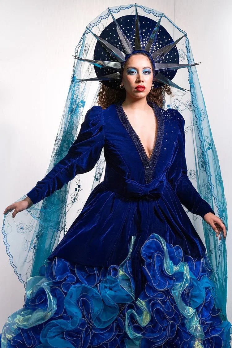 A woman dressed in a vibrant blue velvet gown with ruffled, layered skirt and puffed sleeves. She wears elaborate makeup with blue eyeshadow and red lipstick. Her headpiece is a circular ornate crown with black rays and a lace veil with floral embroi