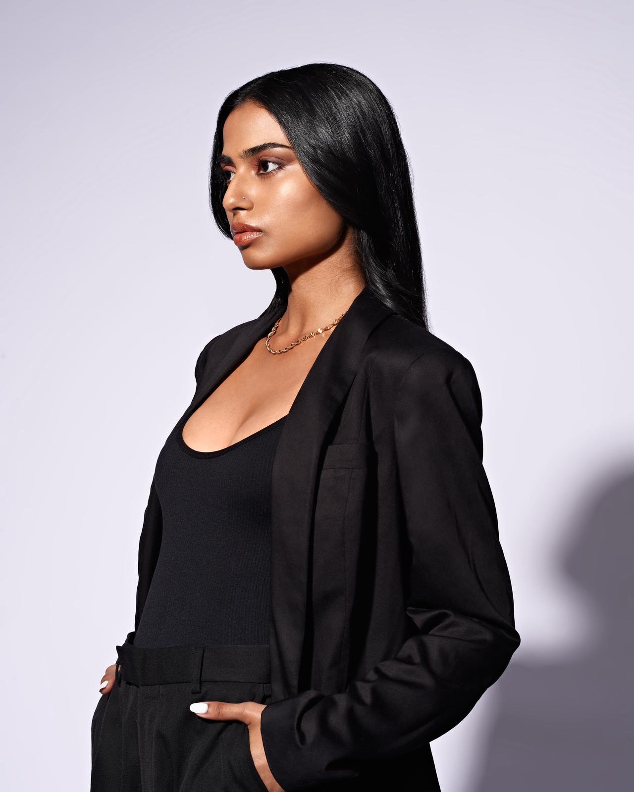 A woman with long black hair, wearing a black blazer, black top, and black pants, standing against a plain light gray background.