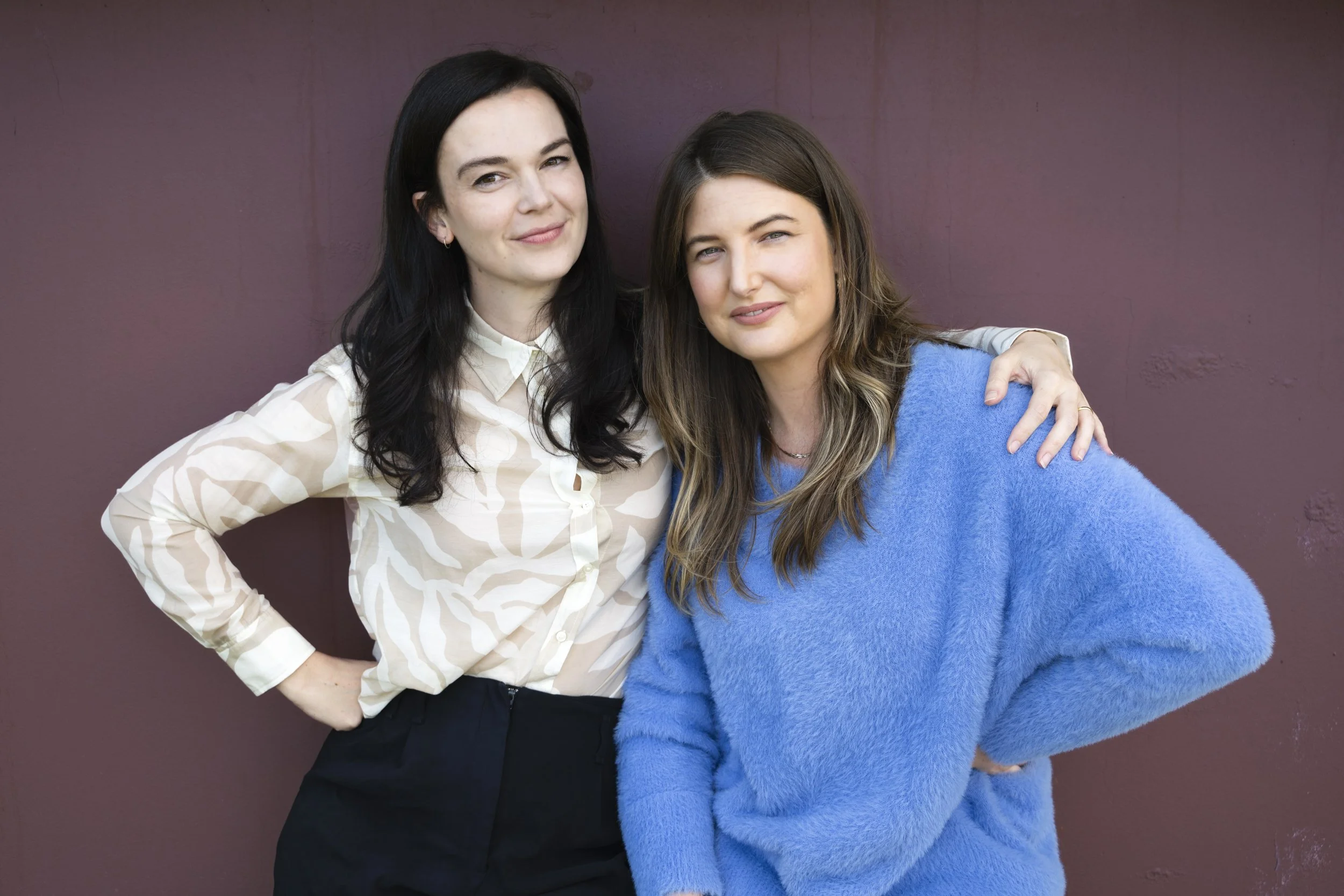 Two women standing closely together against a maroon wall, one with black hair wearing a beige patterned blouse, the other with brown hair wearing a blue sweater, both smiling and looking at the camera.
