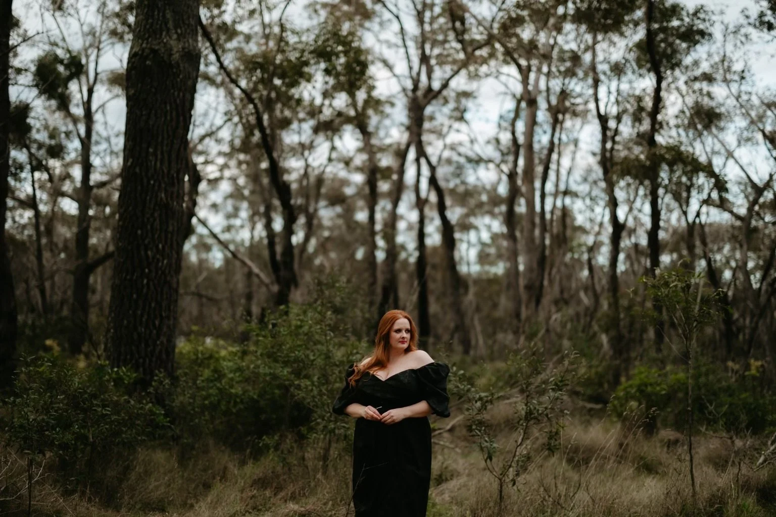 A woman with red hair wearing a black off-shoulder dress standing in a forest with tall trees and green bushes.