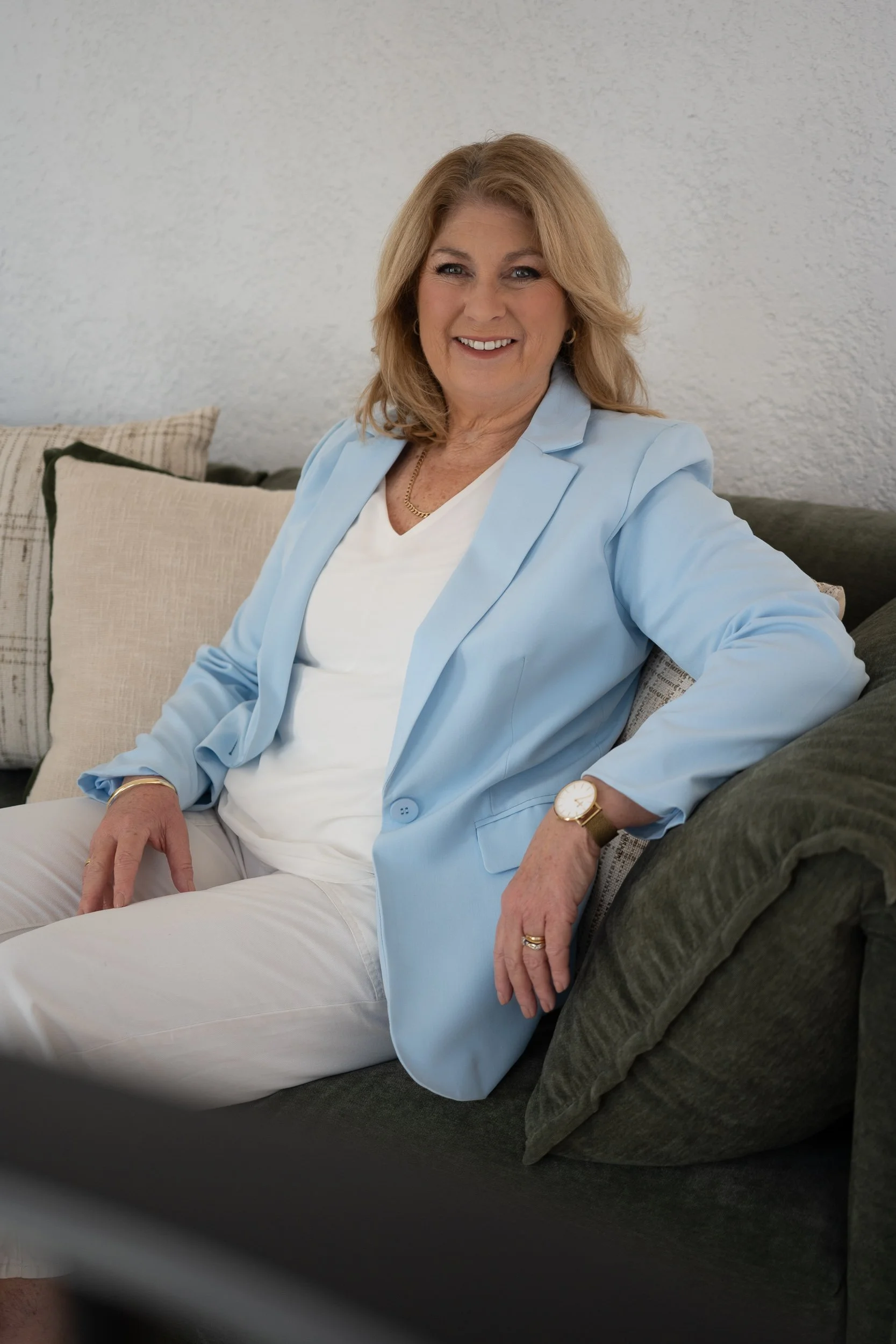 A middle-aged woman with blonde hair, wearing a light blue blazer, white top, and white pants, sitting on a dark green sofa with beige and checkered cushions, smiling at the camera.
