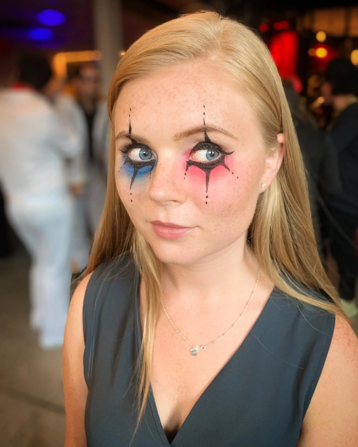 A woman with clown makeup, including blue and pink tears under her eyes, in a crowded indoor setting.