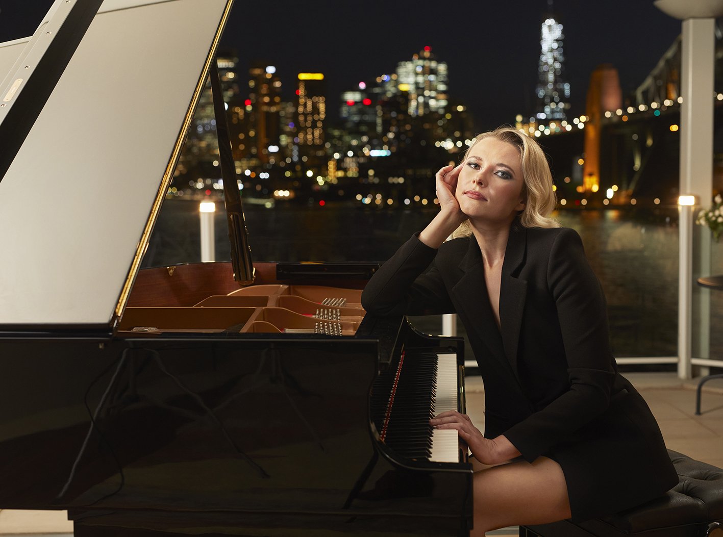 Woman in black blazer sitting at a grand piano with city skyline lights in the background at night.