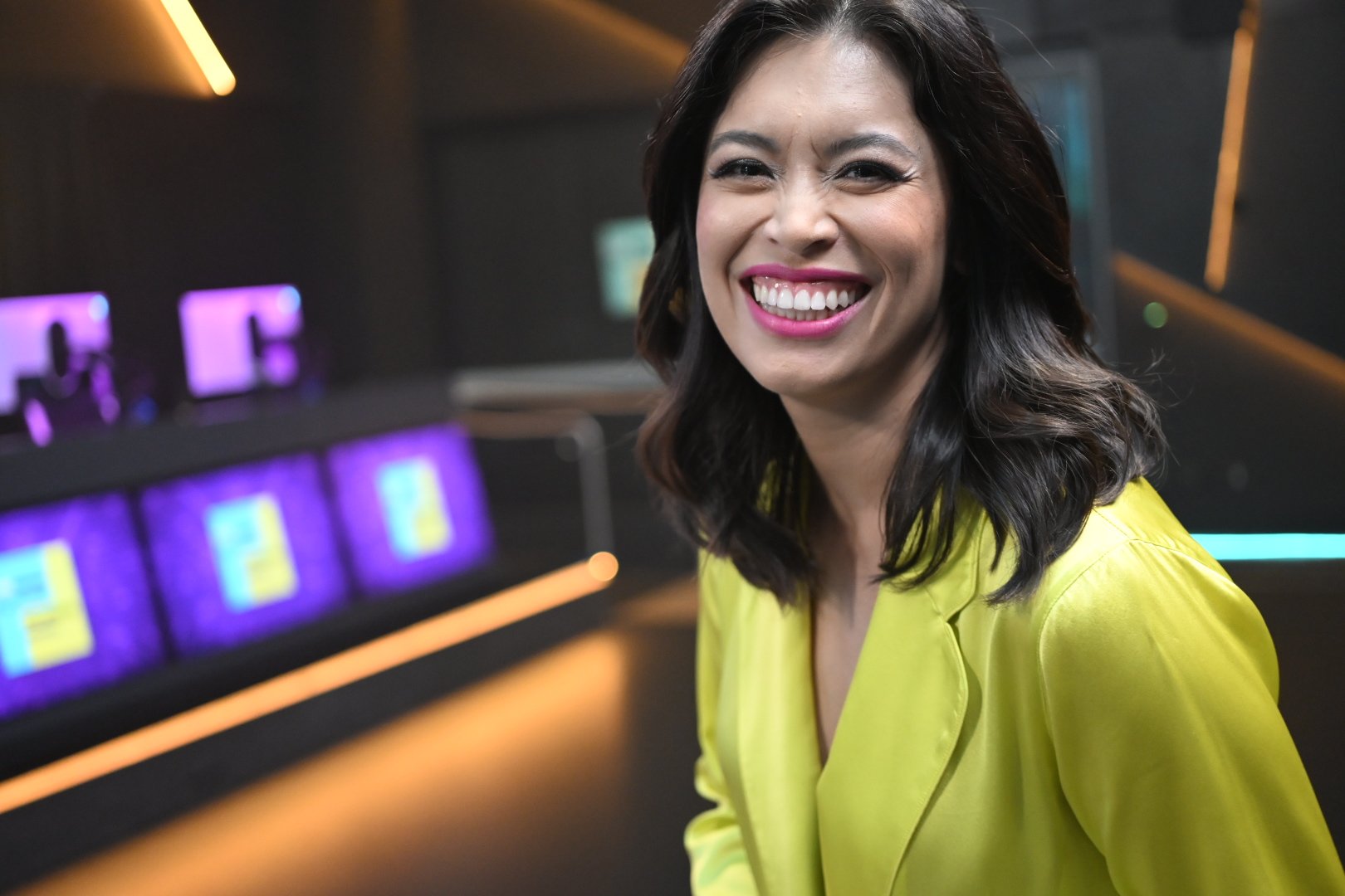A woman with dark hair, wearing a bright yellow blazer, smiling widely in a modern, tech-filled environment with purple and pink illuminated screens in the background.