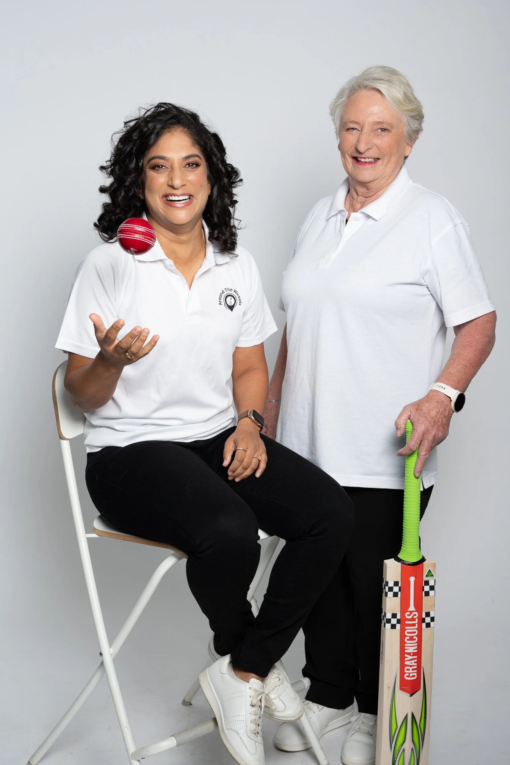 Two women in white shirts with a cricket bat and cricket ball, smiling at the camera against a plain background.