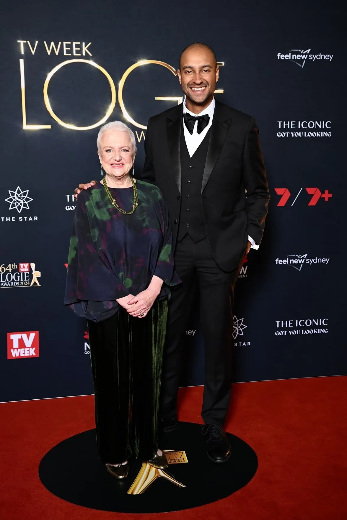 A woman and a man standing together at an awards event in front of a black backdrop with logos and text, smiling for the camera. The woman is wearing a dark floral top with a necklace and dark pants, the man is wearing a black tuxedo with a bow tie a