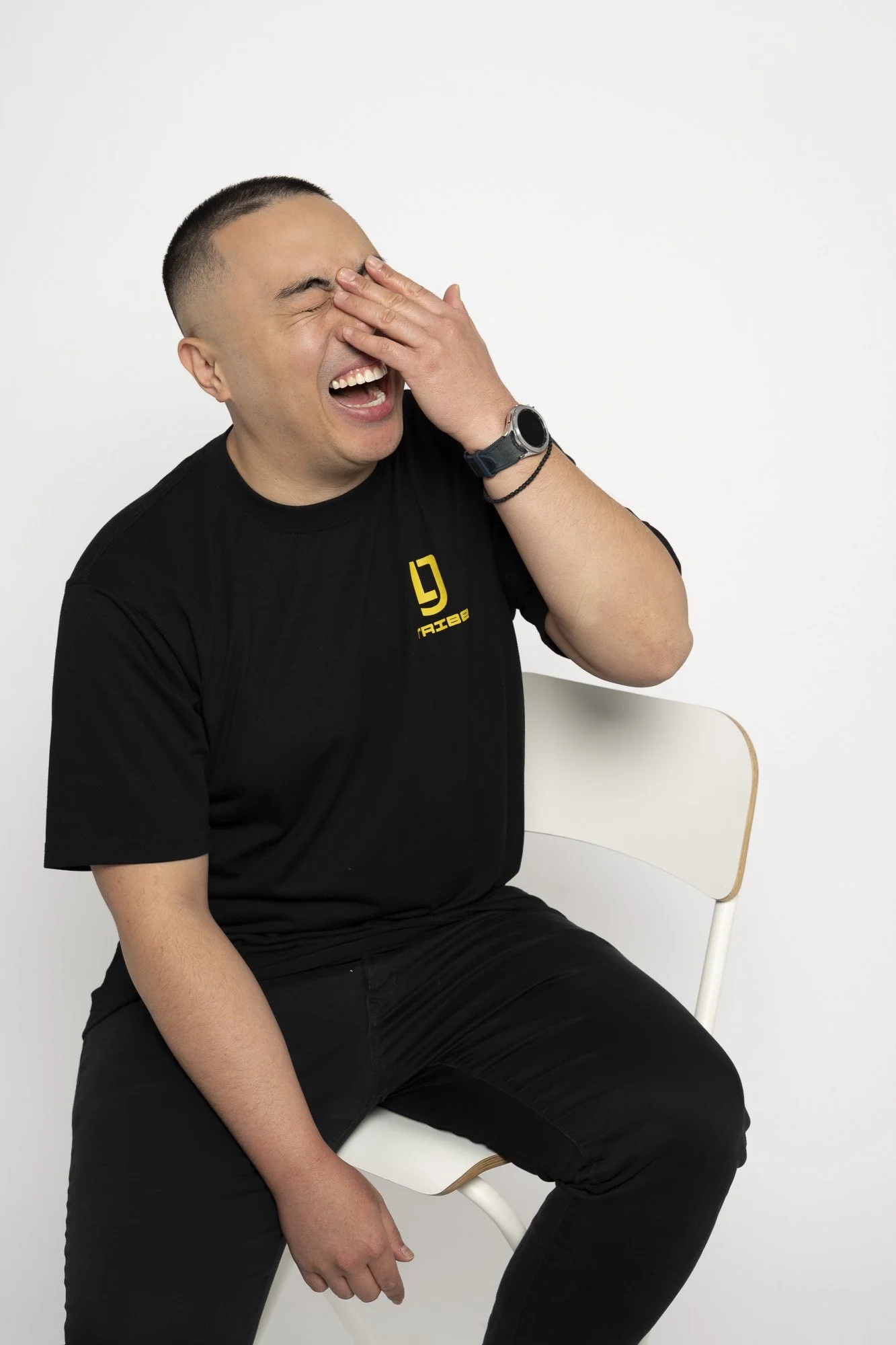A man sitting on a white chair, laughing, covering his face with his right hand, wearing a black t-shirt and black pants, against a white background.