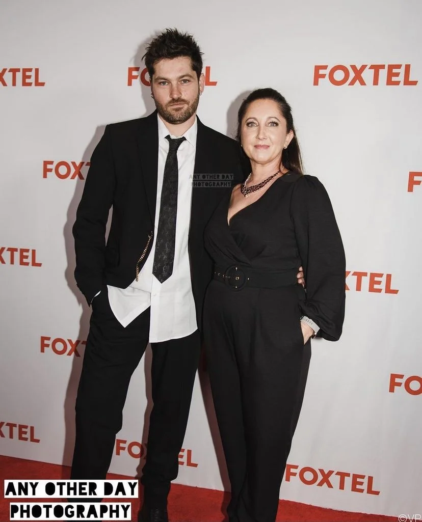 A man and woman standing together on a red carpet at a Foxtel event. The man has dark, spiky hair and a beard, wearing a black suit with a white shirt and a black tie. The woman has dark hair pulled back, wearing a black jumpsuit with long sleeves, a