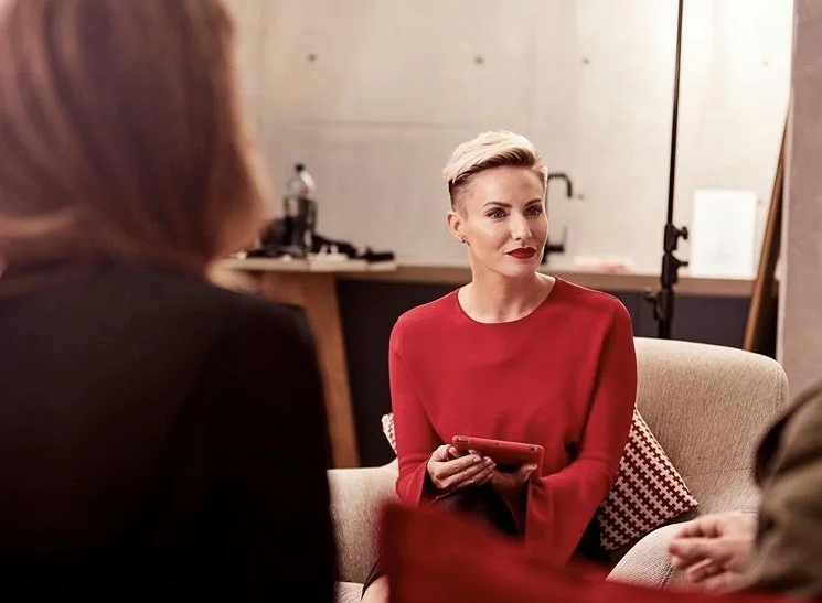 A woman with short platinum blonde hair and red lipstick, wearing a red top, sitting on a beige couch indoors, holding a phone, engaged in conversation with two other people whose faces are not visible.