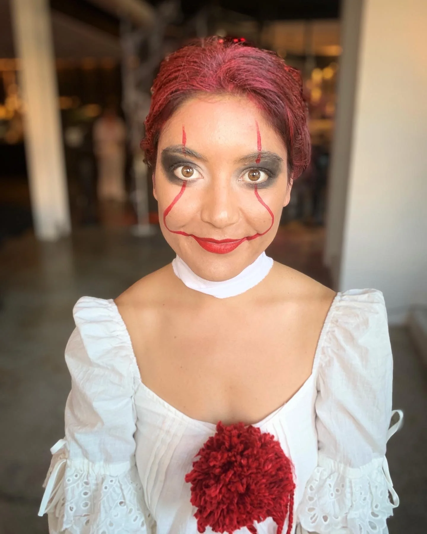 Woman with clown makeup, red wig, white dress, and holding red yarn, standing indoors at night.