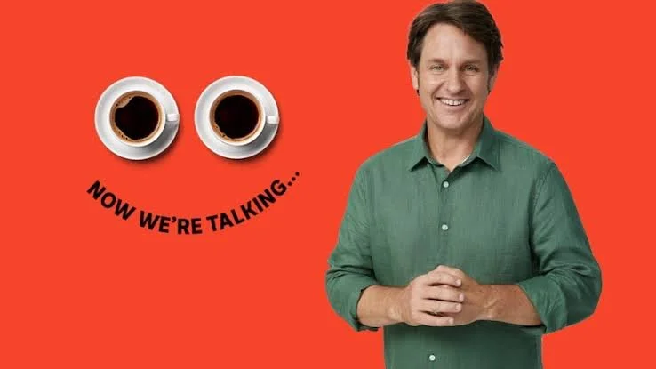 A man with dark hair wearing a green shirt, smiling, with two cups of coffee positioned to look like eyes on a red background, and black text below the cups that reads 'Now we're talking...'