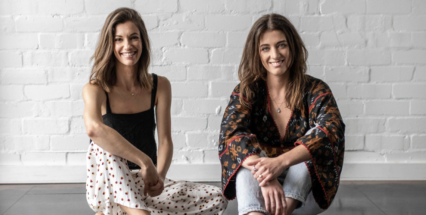 Two young women sitting on the floor in front of a white brick wall, smiling at the camera.