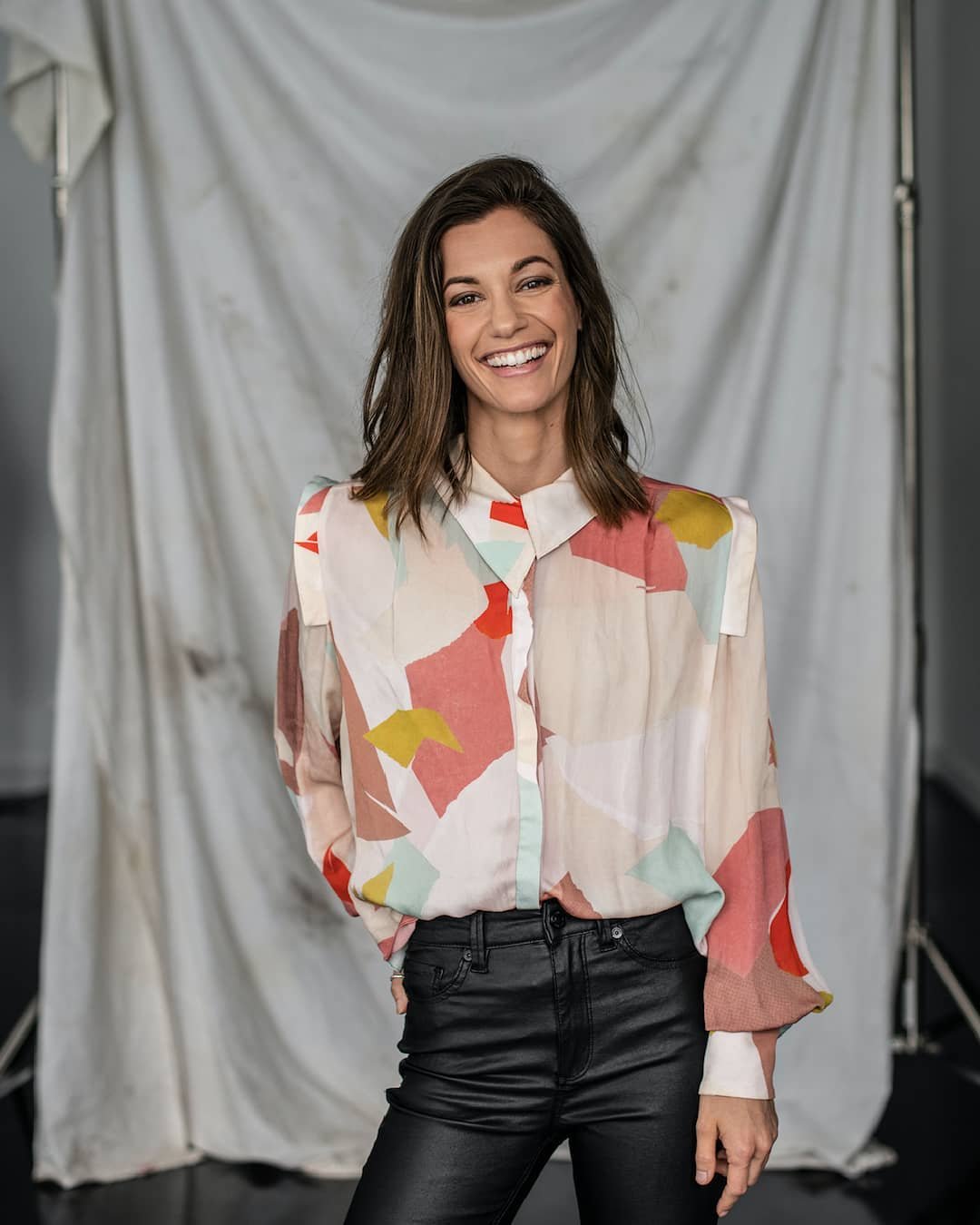 A woman with shoulder-length brown hair smiling, wearing a colorful abstract-patterned blouse and black leather pants, standing in front of a white backdrop.