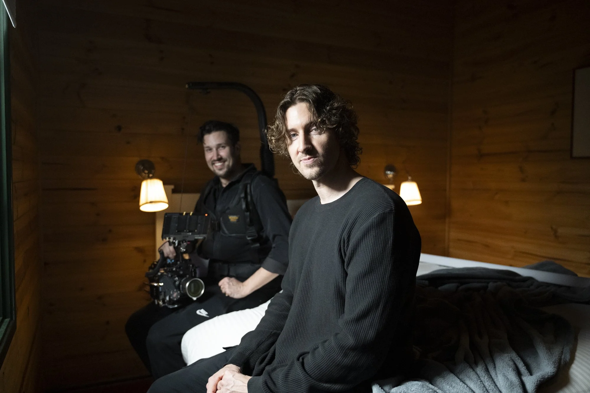 Two men sitting on a bed in a wooden-paneled room, with one holding a camera and the other sitting in front. The room has warm lighting from lamps on the wall. Dean Lewis