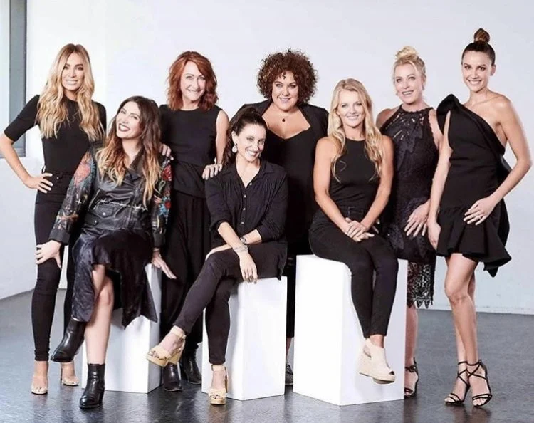 Group of eight women dressed in black, posing together in a studio with a plain white background.