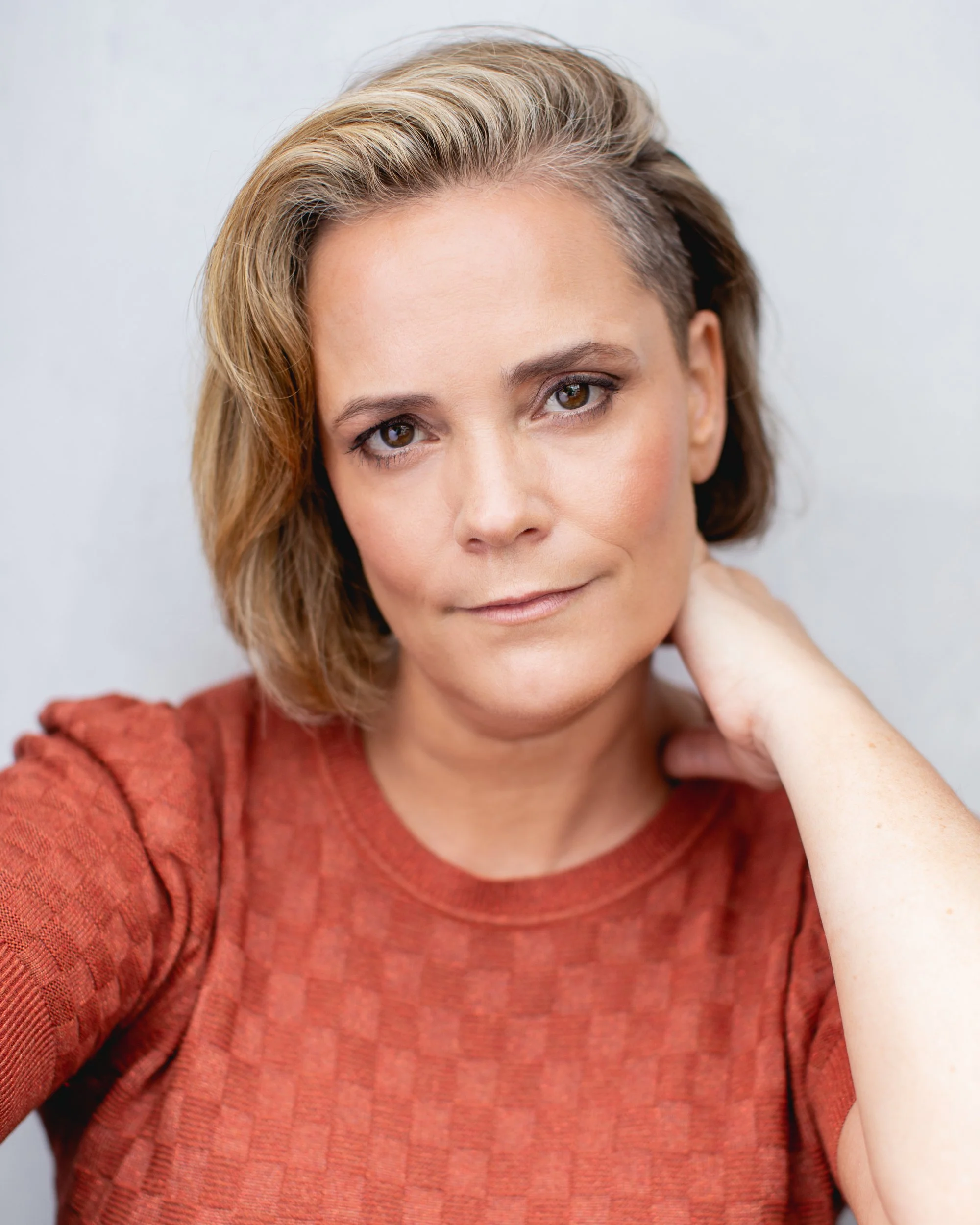A woman with short, styled blonde hair wearing a rust-colored textured top, resting her hand on her neck, looking at the camera.