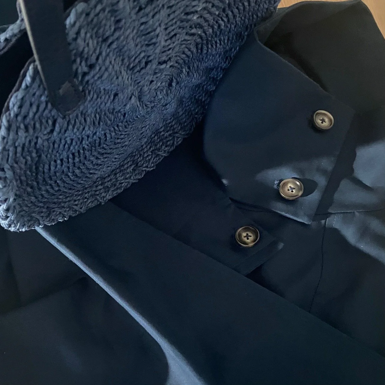 Navy straw bag with structured handle placed against a deep navy tailored coat with silver button details, demonstrating tonal minimalism through texture contrast