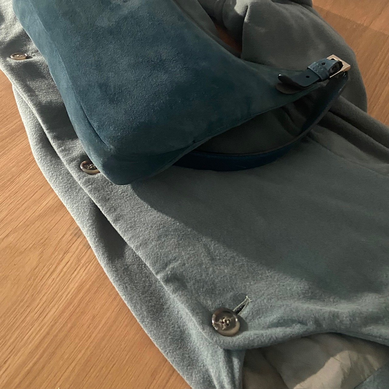 Teal suede bag with silver hardware placed on a folded sage green coat, with a designer label visible, demonstrating luxury tonal dressing in a flat lay composition