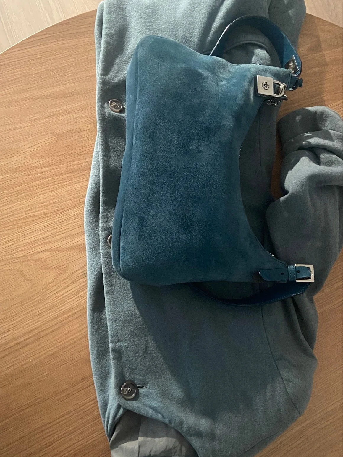 Teal suede slouchy bag resting on a sage green wool coat laid flat on a warm wood surface, showing tonal colour coordination between coat and bag