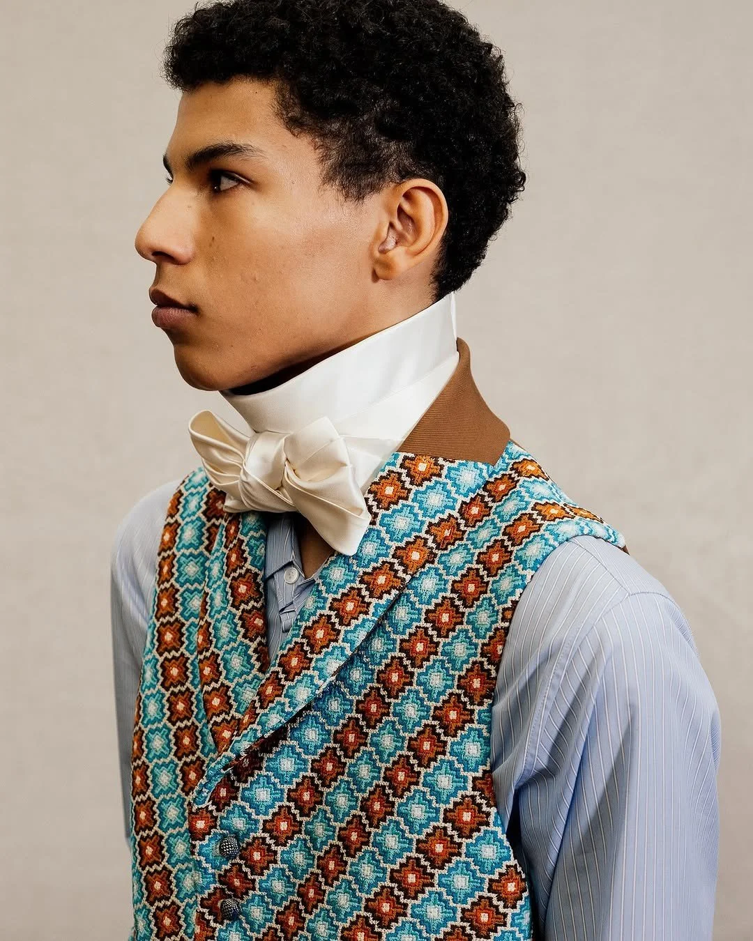 Close-up of a look from Jonathan Anderson’s debut menswear collection featuring a crisp striped shirt, bright patterned woven vest, a silk bowtie, and a high-neck collar.