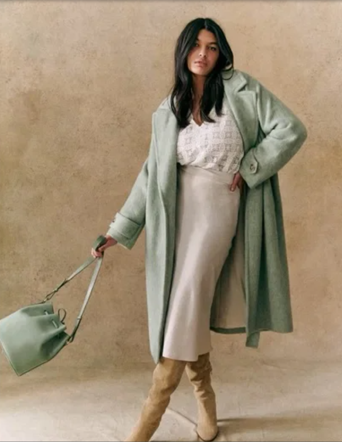 Model wearing an oversized sage green wool coat and carrying a matching sage green bucket bag against a neutral background, illustrating tonal minimalism as a complete luxury outfit