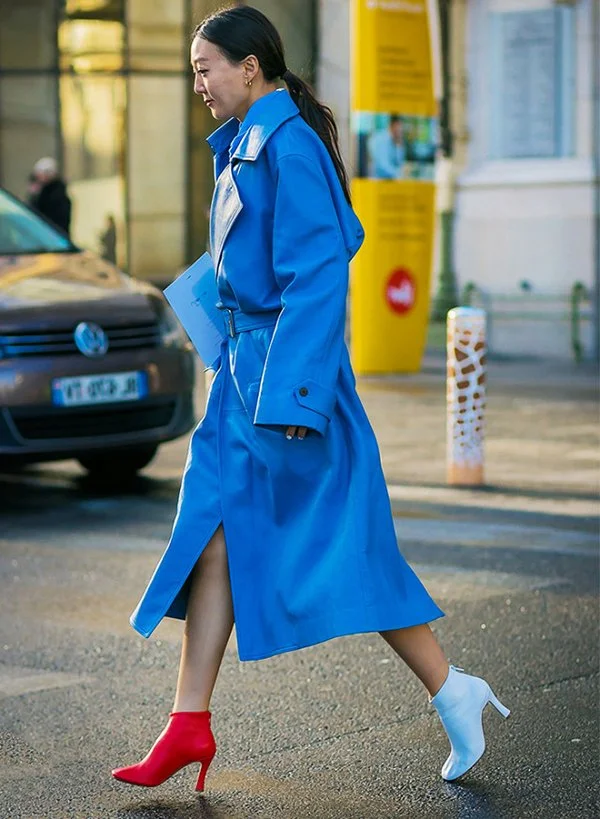 A woman walks across the street in colourful mismatched heeled boots and coordinating bold trench coat, emphasizing asymmetry and risk.