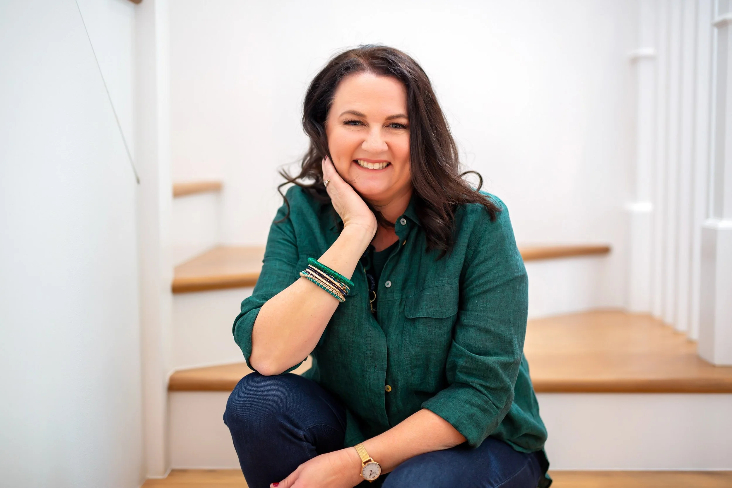 Trina Julius - Red Cherry Photography Education -   shoulder-length dark hair, wearing a green button-up shirt, sitting on wooden stairs indoors, smiling and resting her chin on her hand.
