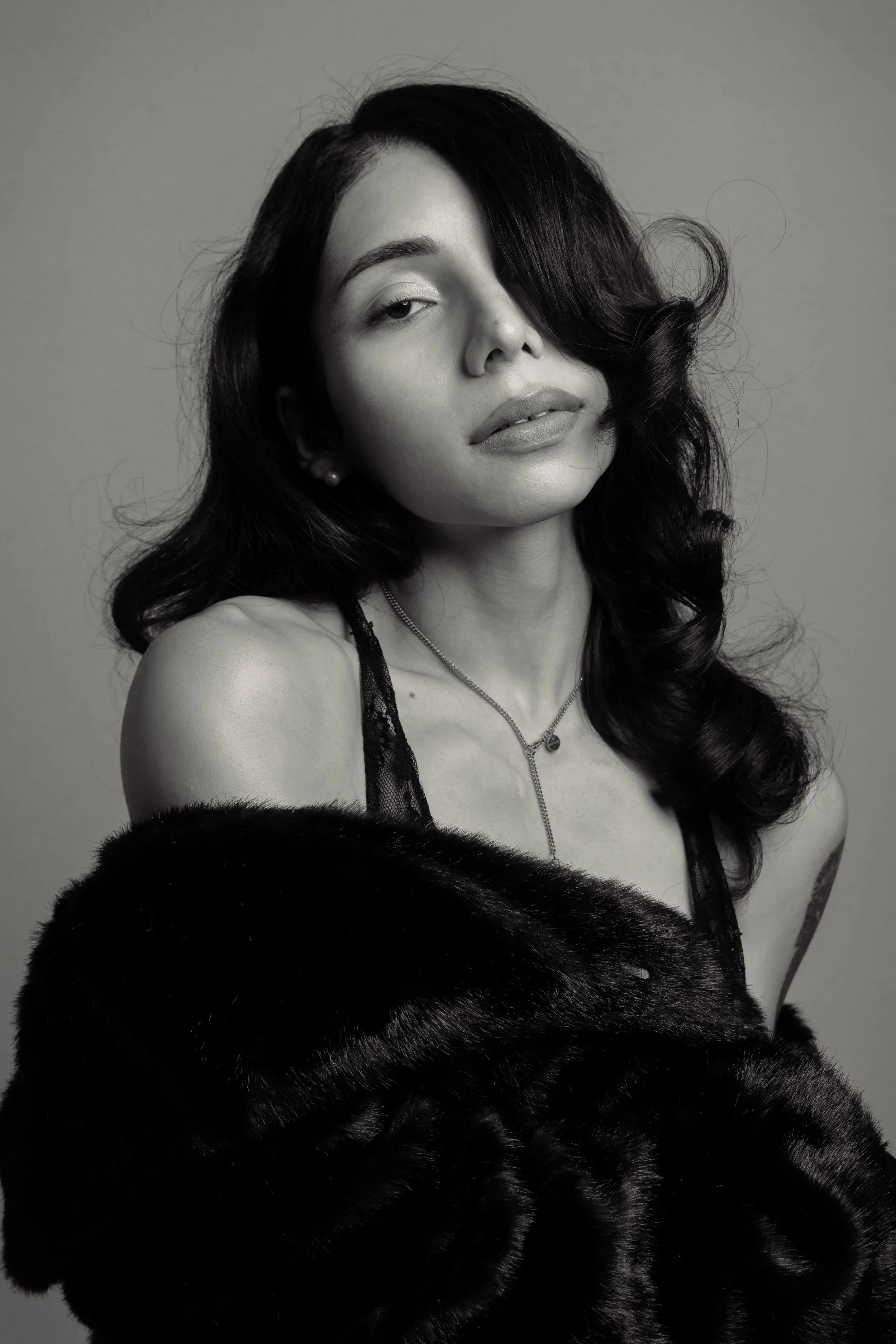 Black and white editorial portrait of actress Yasha la Bella in THE STARLET photo story, an exclusive collaboration with Emcee studios. She wears a black lace Collina Strada dress and a black fur coat, with her long wave hair covering one eye.