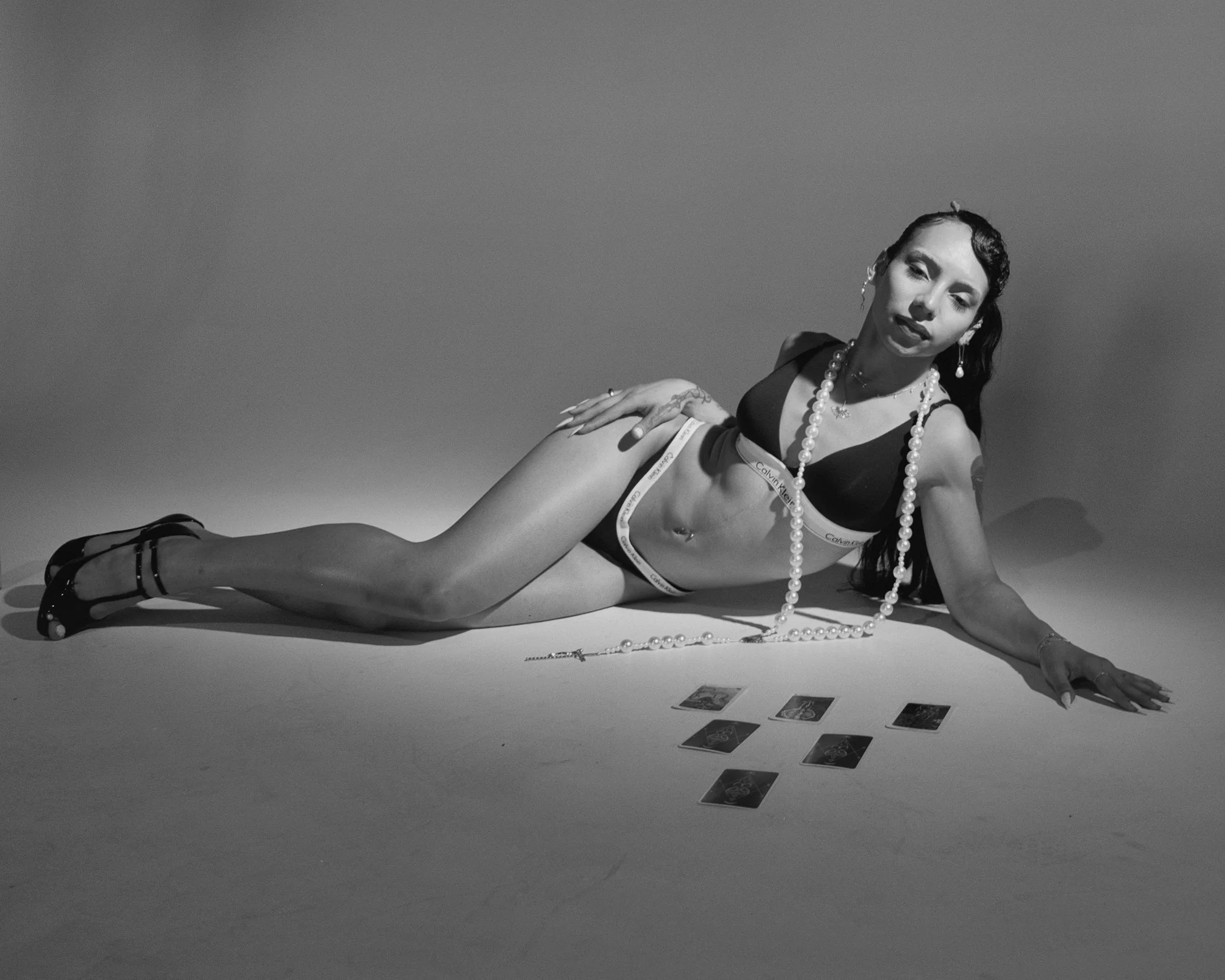 A woman lying on her side on the floor, dressed in black lingerie, high heels, and wearing jewelry, with tarot cards spread out in front of her.