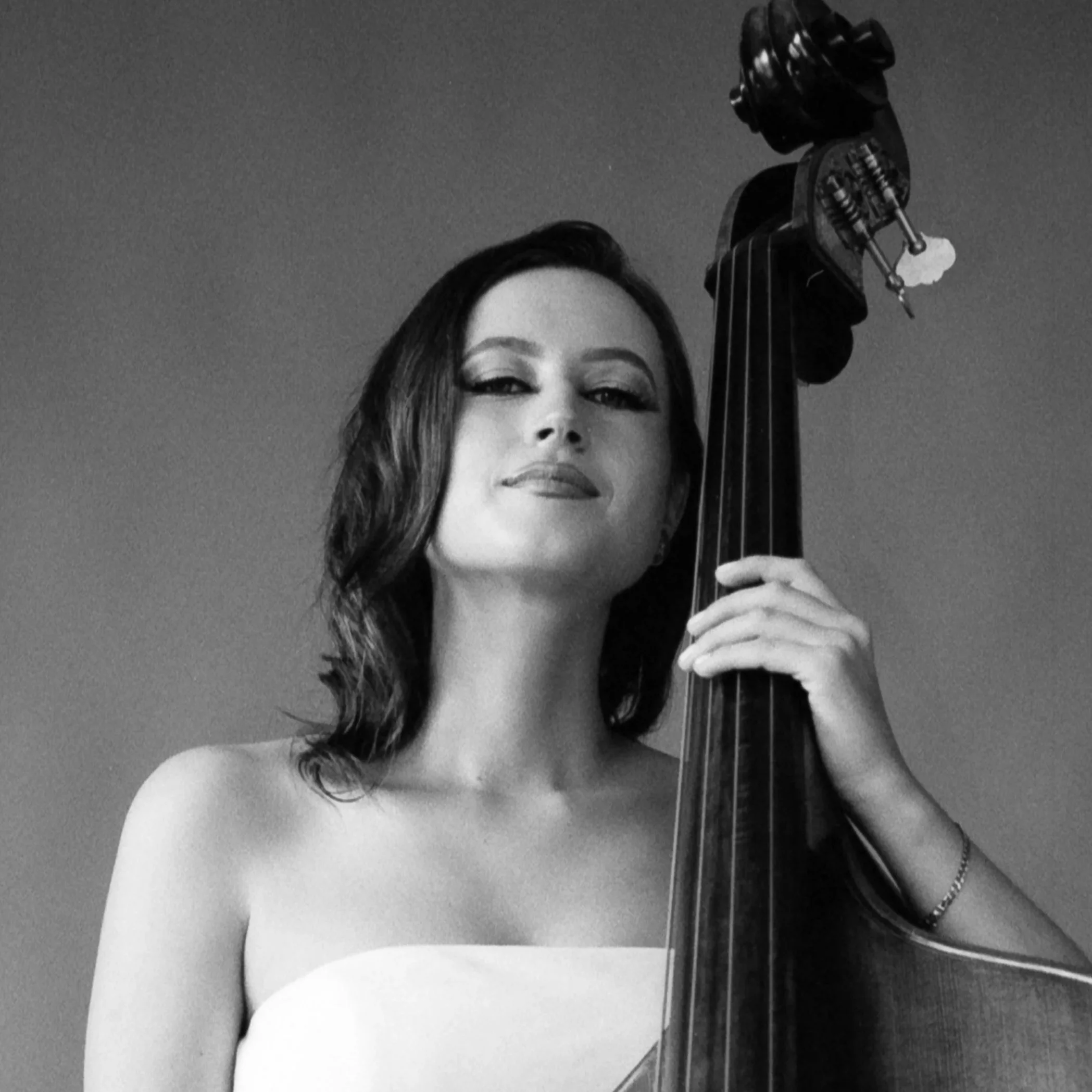 A black and white photo of a woman with long hair holding a cello, with her face partially visible and a slight smile.