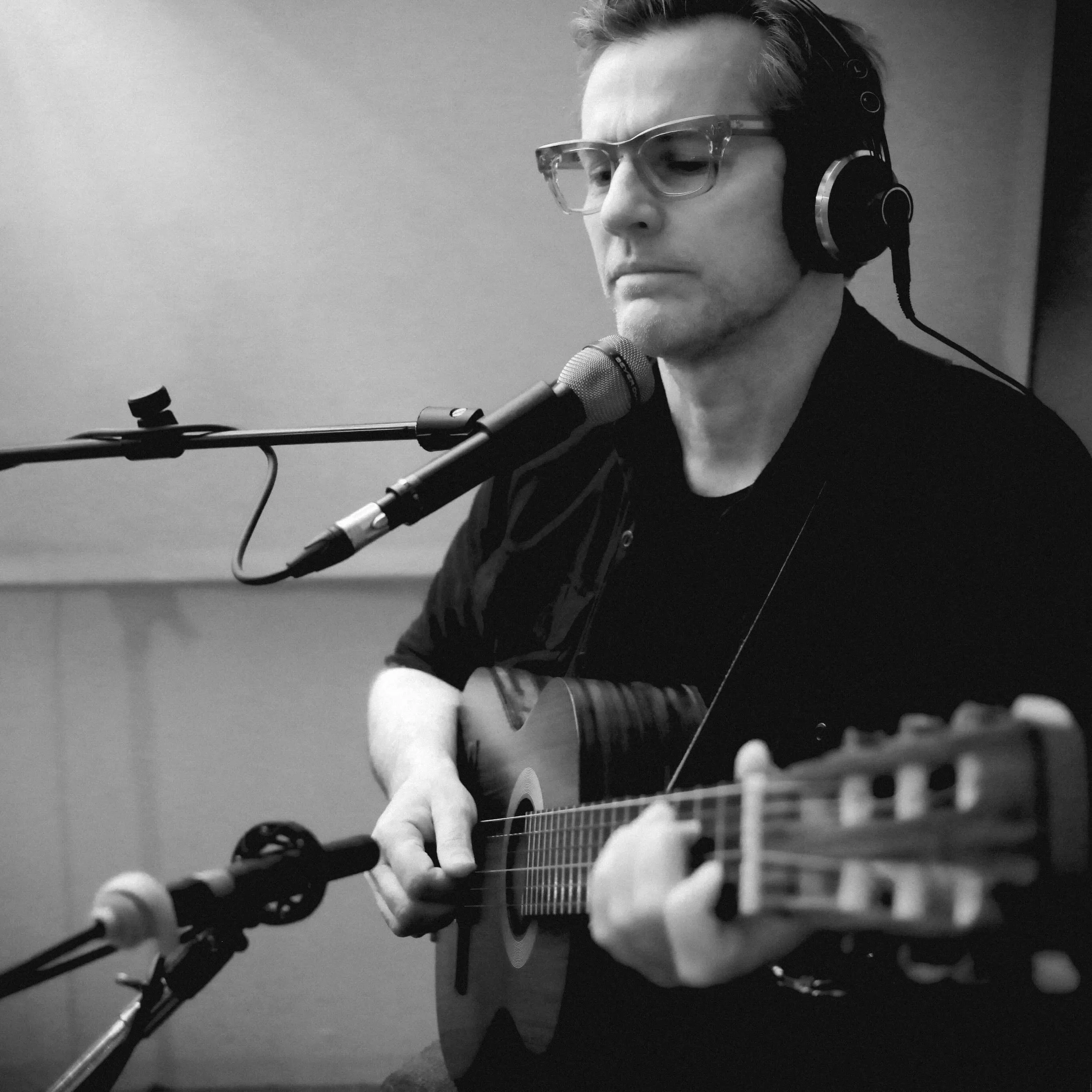 A person wearing glasses and headphones is playing an acoustic guitar into a microphone in a recording studio.