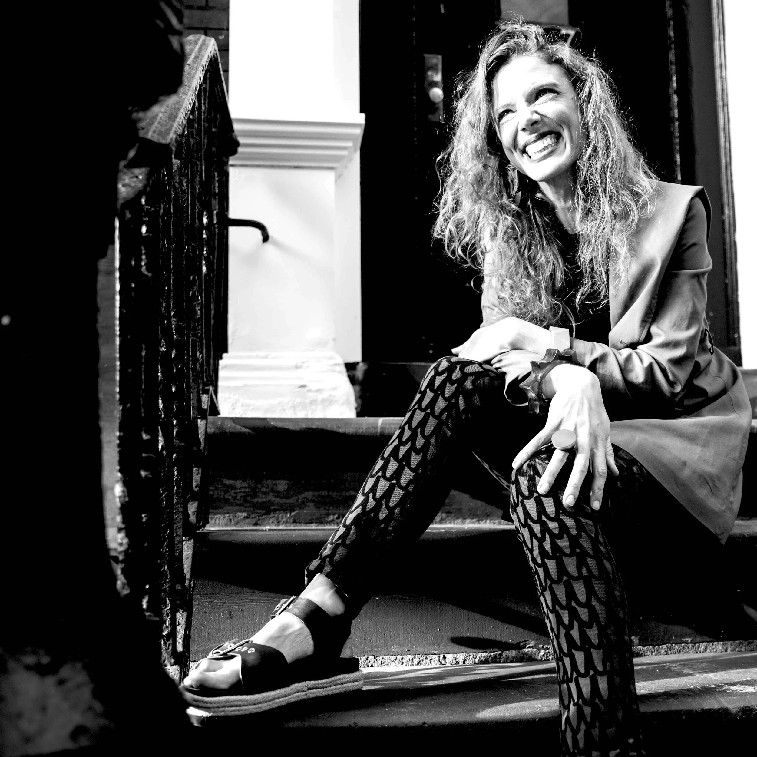 Australia's leading Jazz vocalist, smiling and squinting, sitting on a NY stoop beside a door with a black and white patterned dress and a jacket draped over her lap.