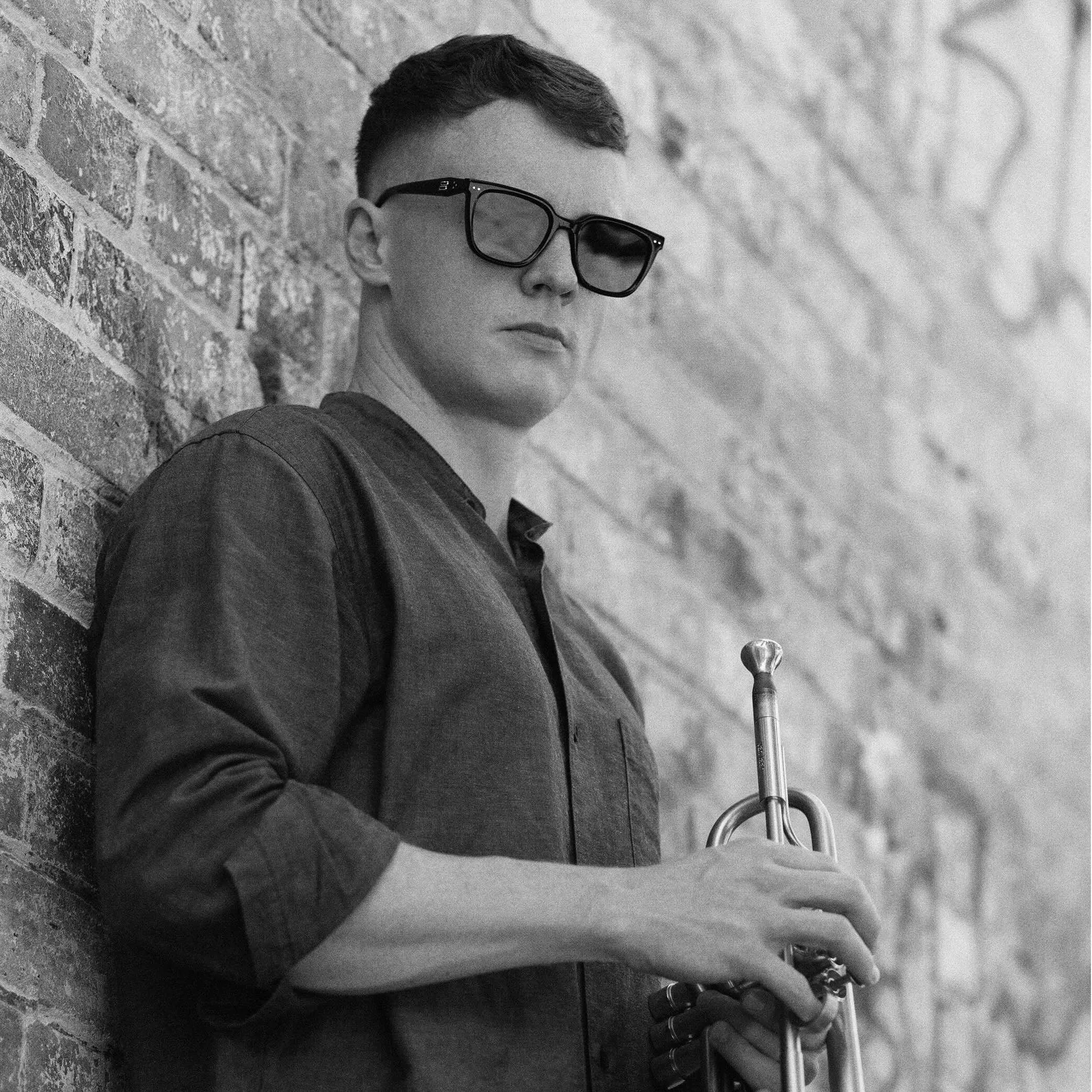 Black and white photo of a award winning jazz trumpeter with short hair wearing sunglasses, leaning against a brick wall, holding a skateboard.
