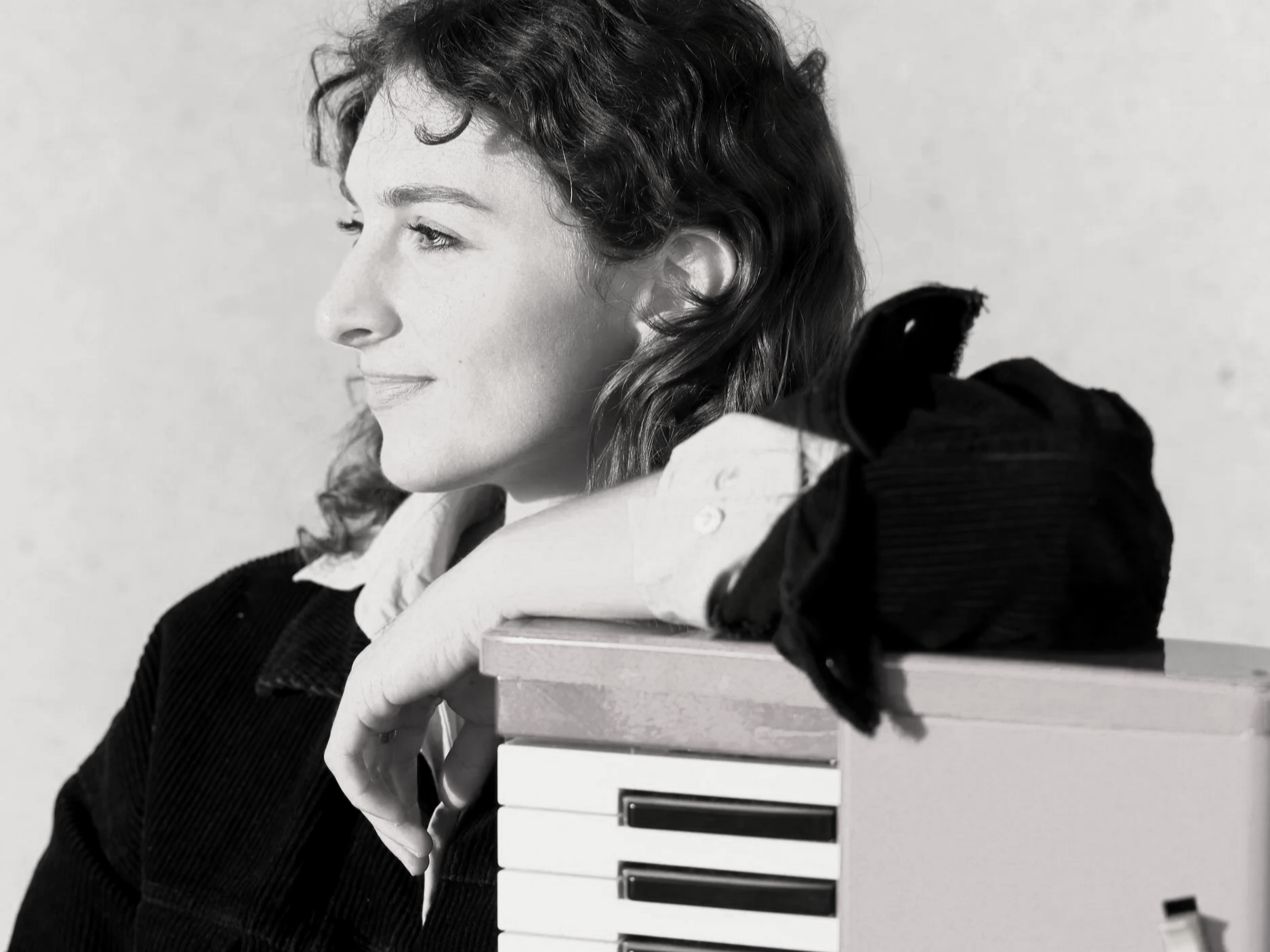 A hip female musician with wavy hair looking to the side, resting her chin on her arm, which is on a stack of books or boxes. The image is in black and white.