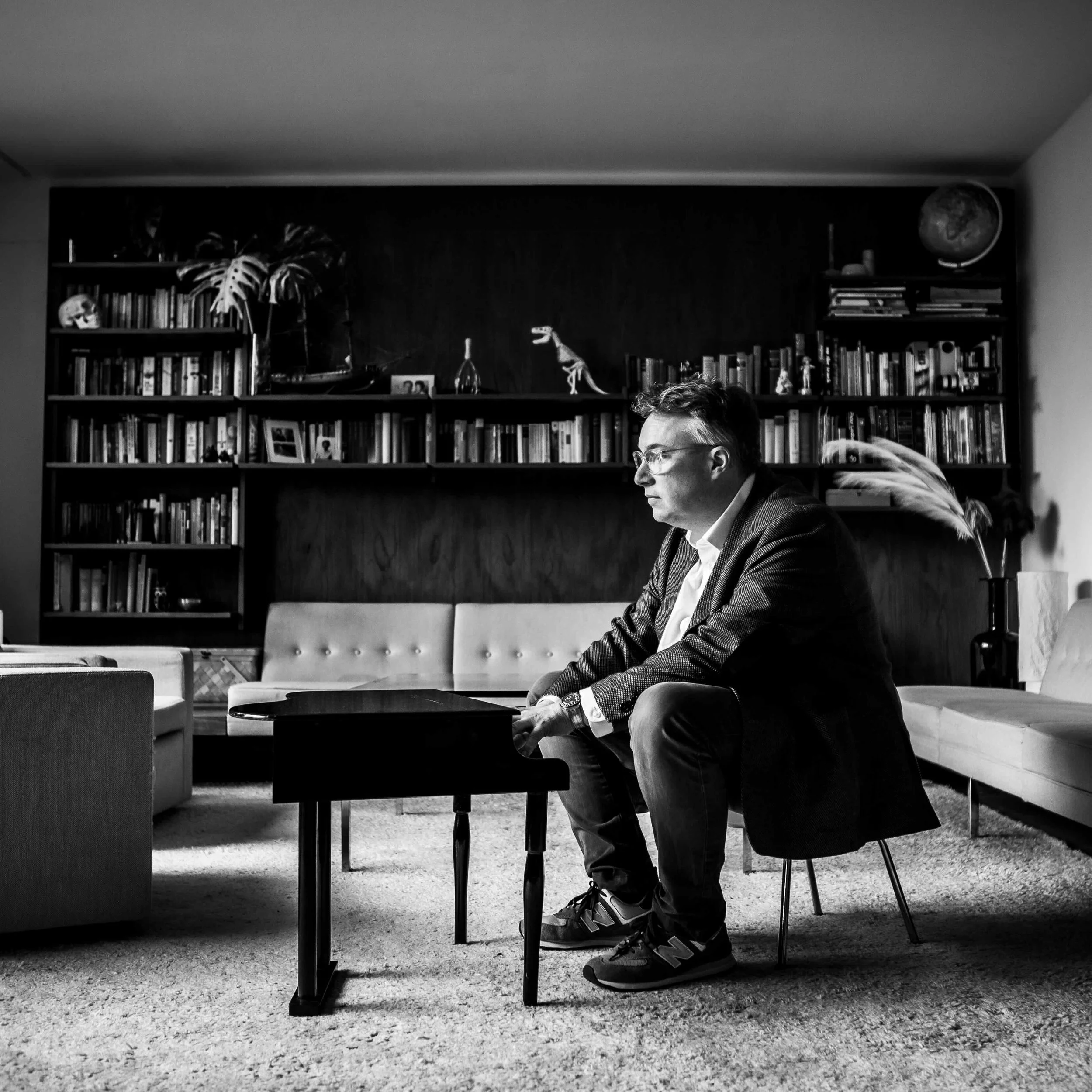 Black and white photo of a man sitting at a table in a living room, with bookshelves and decorative items, including a globe and model dinosaur in the background, and a woman with her arm around him.