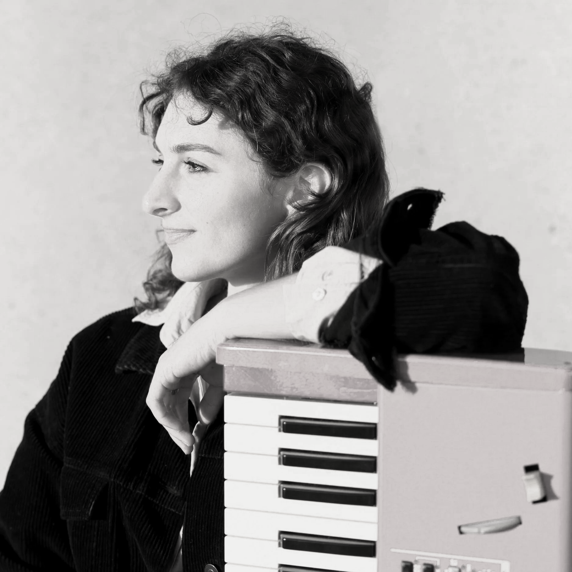 Black and white photo of a woman with curly hair leaning on a piano, her hand on her chin, appearing contemplative.